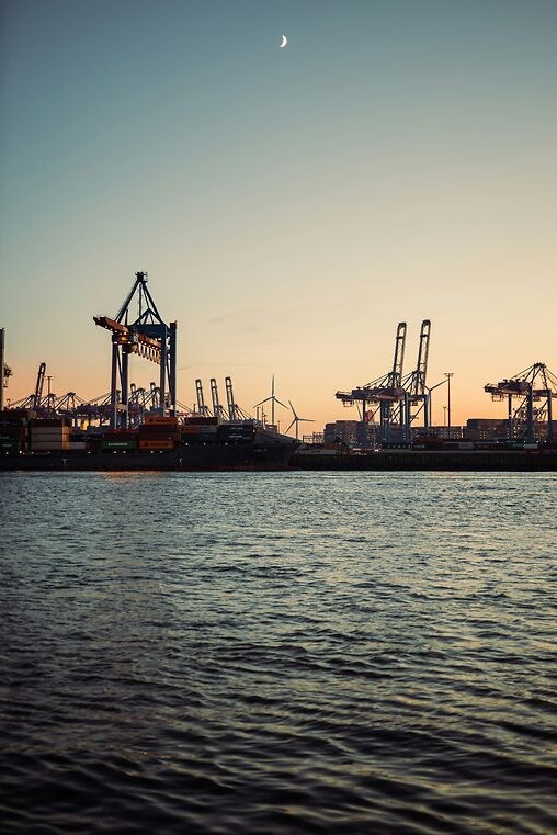 Blick auf Containerkräne und Frachtschiffe im Hamburger Hafen bei Sonnenuntergang – maritimes Panorama über der Elbe.